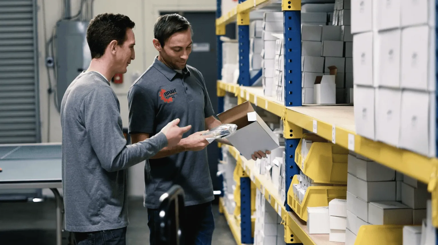 Two BRAH Electric team members checking a packaged part beside warehouse shelving